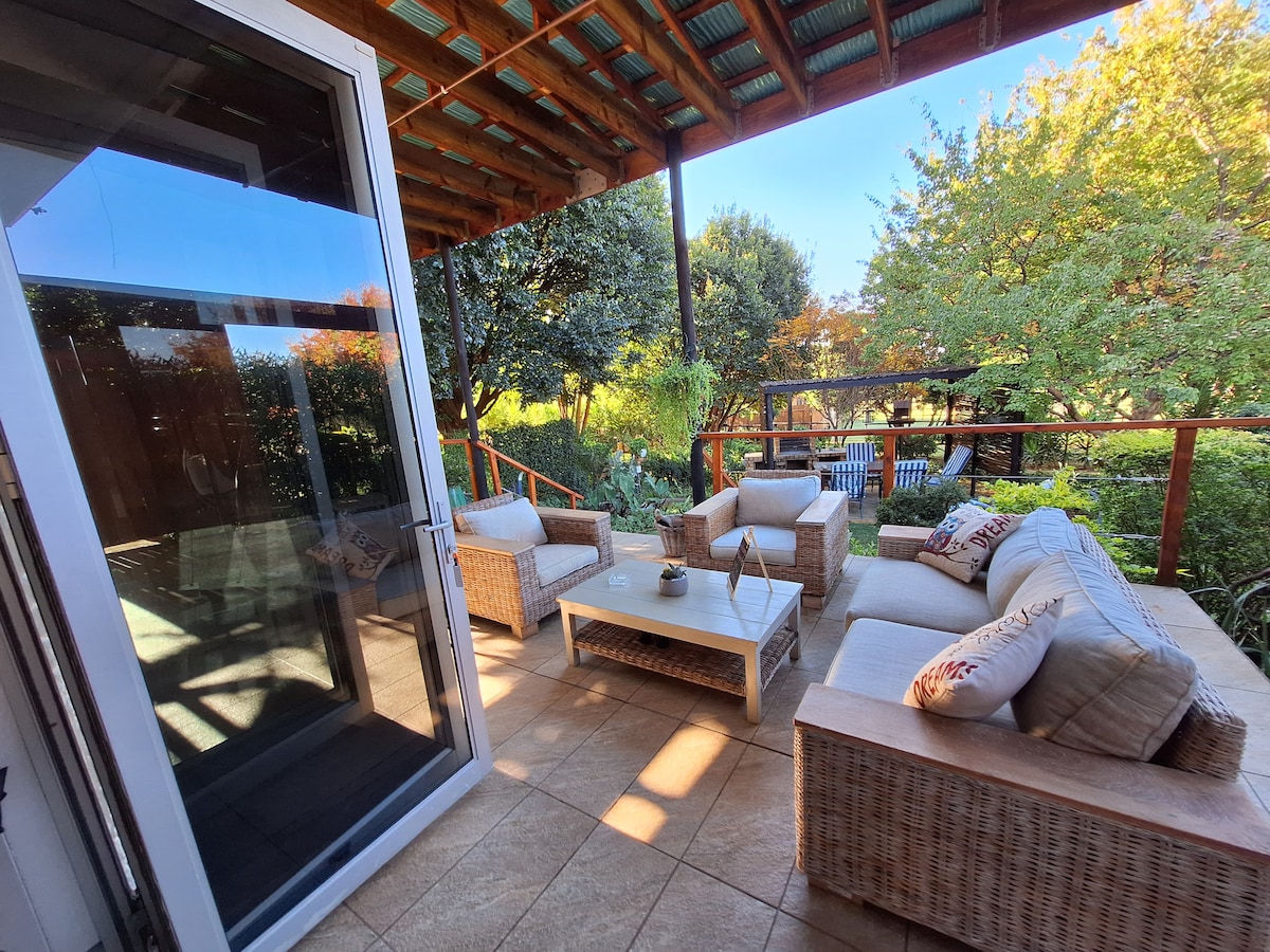 An outdoor seating area features a light-colored wicker sofa and chairs arranged around a low coffee table. Sunlight filters through trees, casting soft shadows on the tiled floor, while the lush garden beyond adds greenery to the peaceful environment.