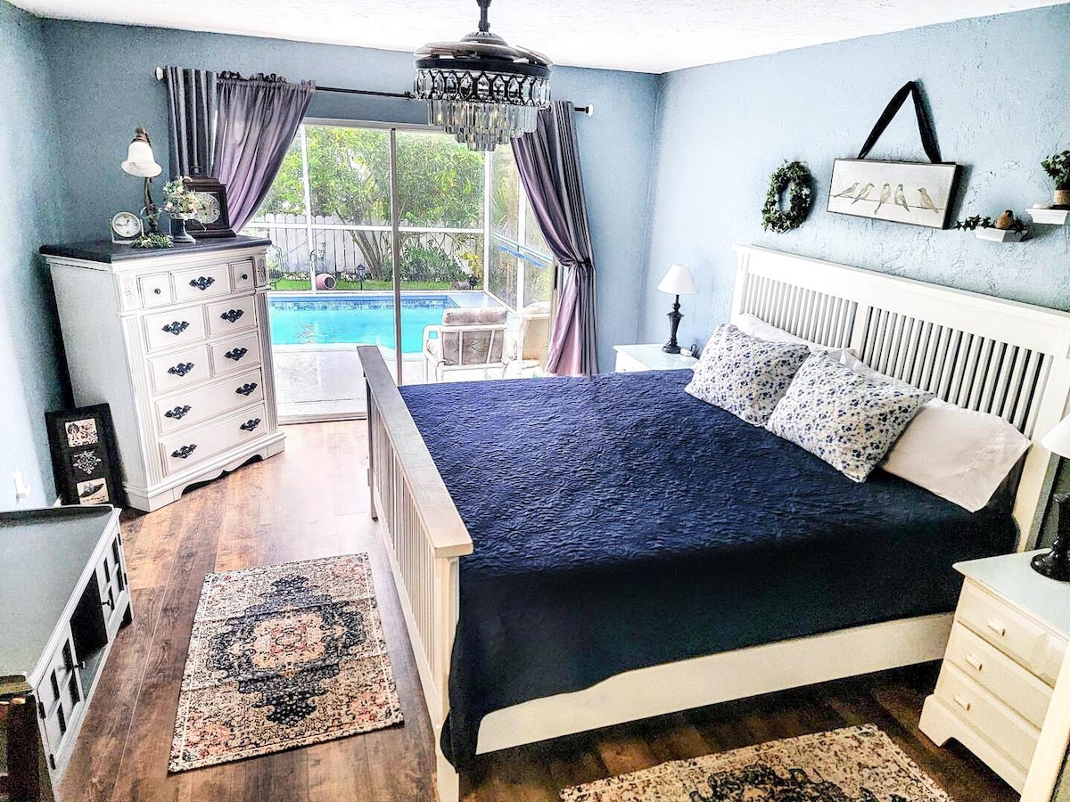 A spacious bedroom features a king bed with a dark blue coverlet, complemented by patterned pillows. Natural light flows in through sliding glass doors, revealing a view of the outdoor pool area. A dresser and side tables provide functional storage and surface space.
