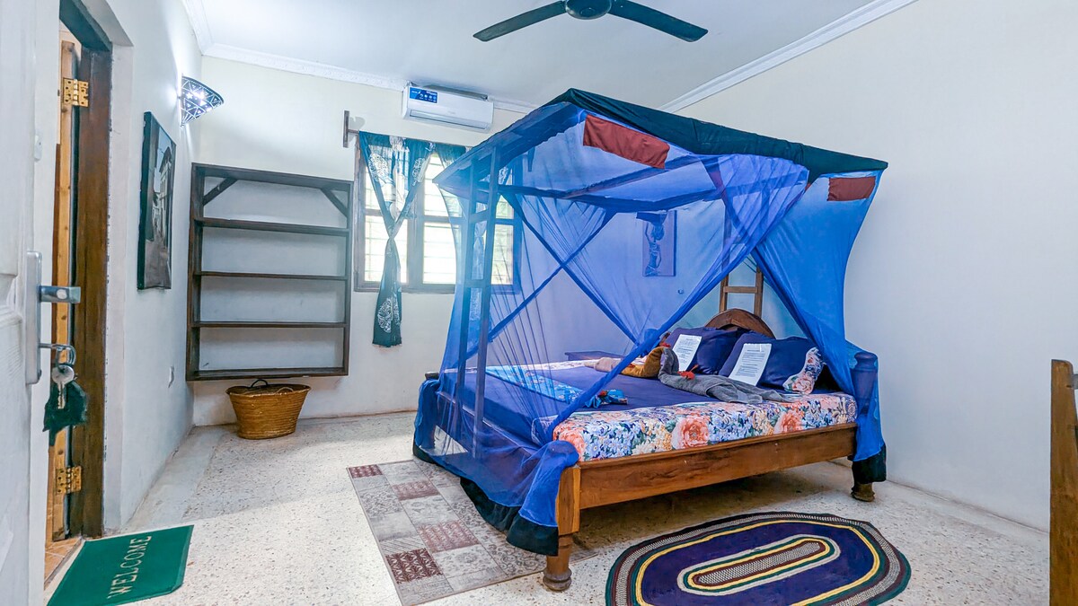 A spacious bedroom features a large bed draped with a netting canopy, providing a serene sleeping area. Shelves are positioned along the walls for storage, and a woven basket adds a natural touch. Soft light filters through the curtain-covered windows, illuminating the calming colors of the decor.