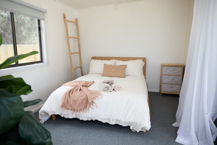 The "Out House" bedroom features a double bed facing the ocean. A minimalist design with soft linens, a ladder for decor, and a leafy potted plant adds a cozy and peaceful touch to the space. Perfect for a serene, ocean-facing retreat.