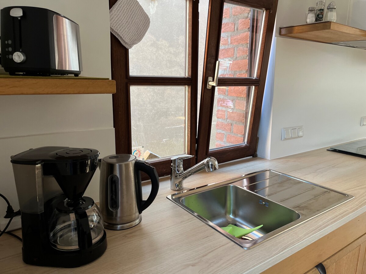 A well-equipped kitchen features modern appliances including a coffee maker, kettle, and a stainless-steel sink. Sunlight filters through the window above the sink, illuminating the wooden countertop and creating a functional cooking space.