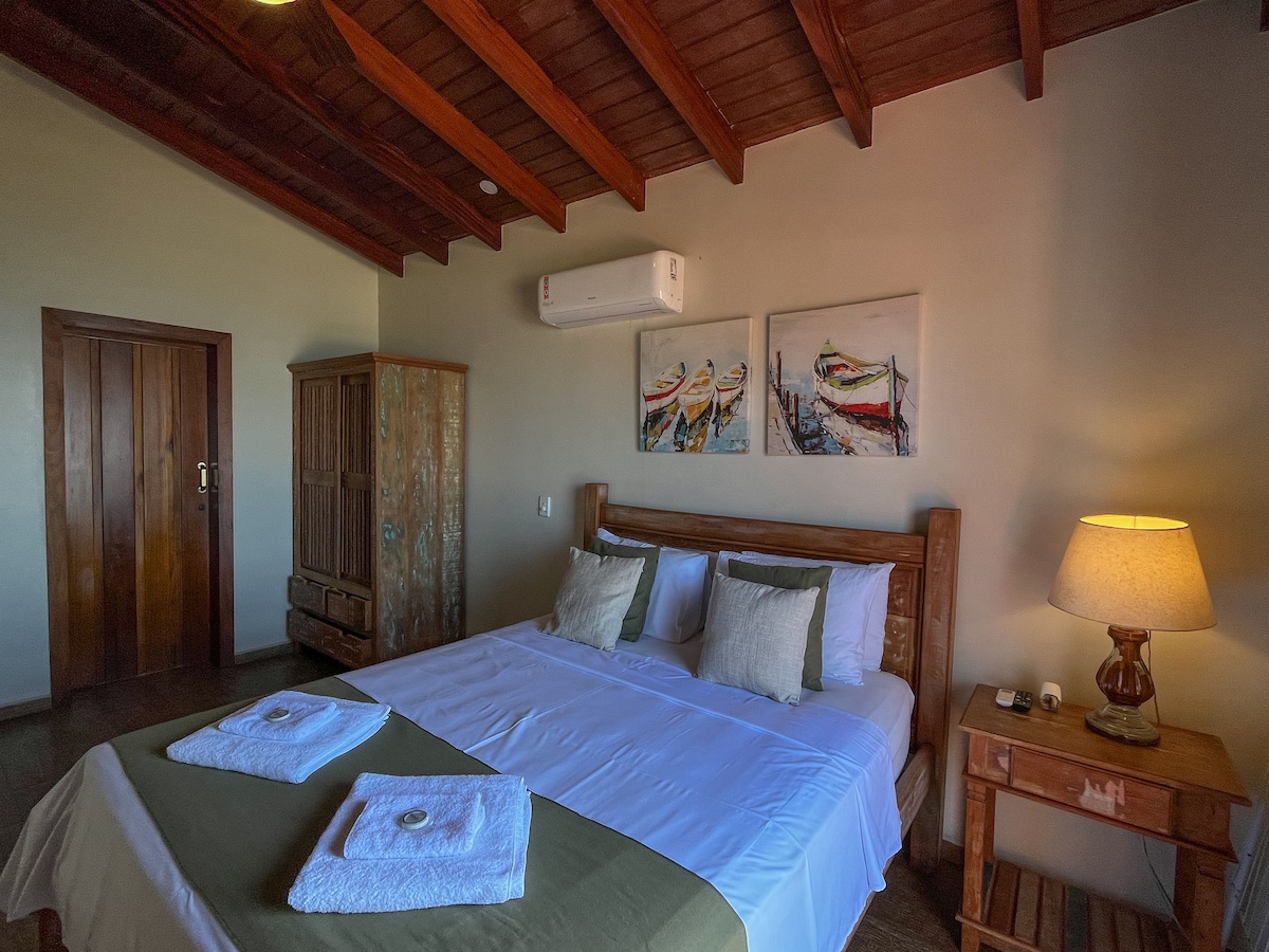 The spacious bedroom features a wooden ceiling and is furnished with a large bed adorned with decorative pillows. A bedside table holds a lamp, and artwork depicting colorful boats is displayed on the wall. A wooden wardrobe adds to the room's warm aesthetic.