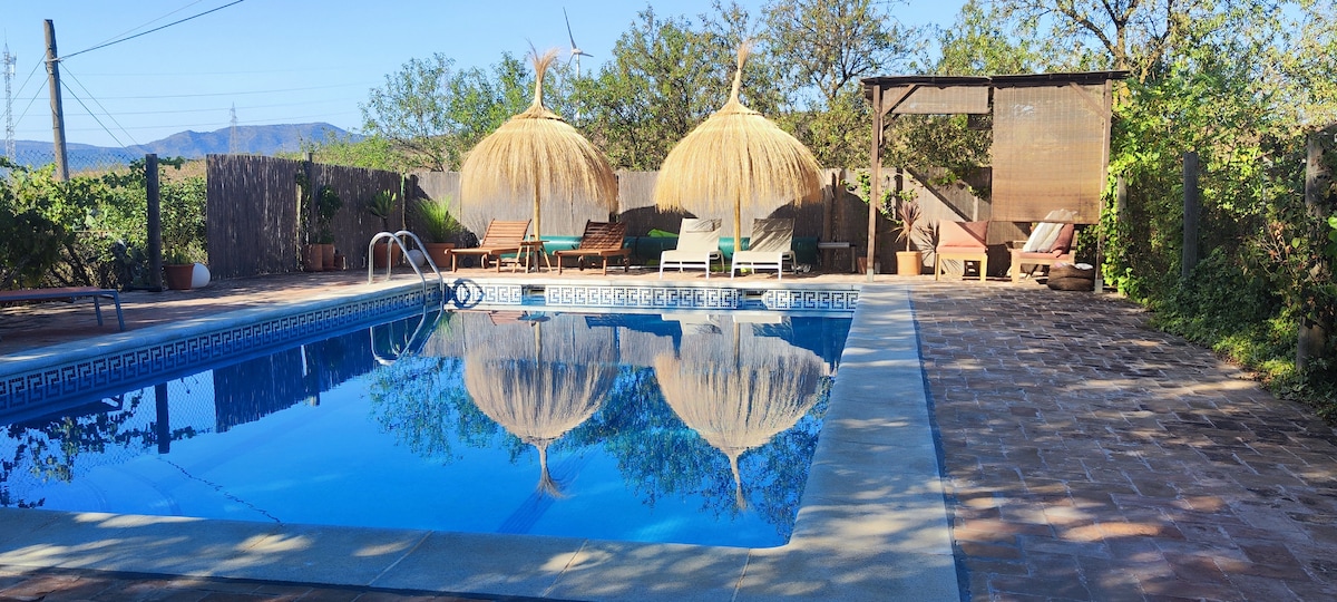 A serene outdoor pool area is surrounded by lush greenery and features two thatched umbrellas offering shade. Sun loungers are positioned nearby, inviting relaxation by the crystal-clear water. The tiled pool deck provides ample space for enjoying the tranquil landscape.
