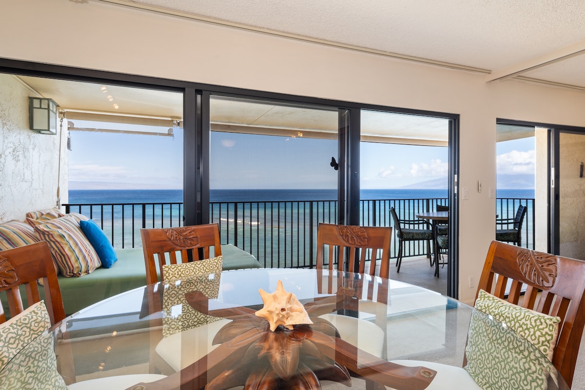 A dining area with a round glass table is surrounded by wooden chairs, offering seating for six. Large sliding glass doors open to a lanai, showcasing expansive ocean views and outdoor seating. A decorative starfish rests at the center of the table.