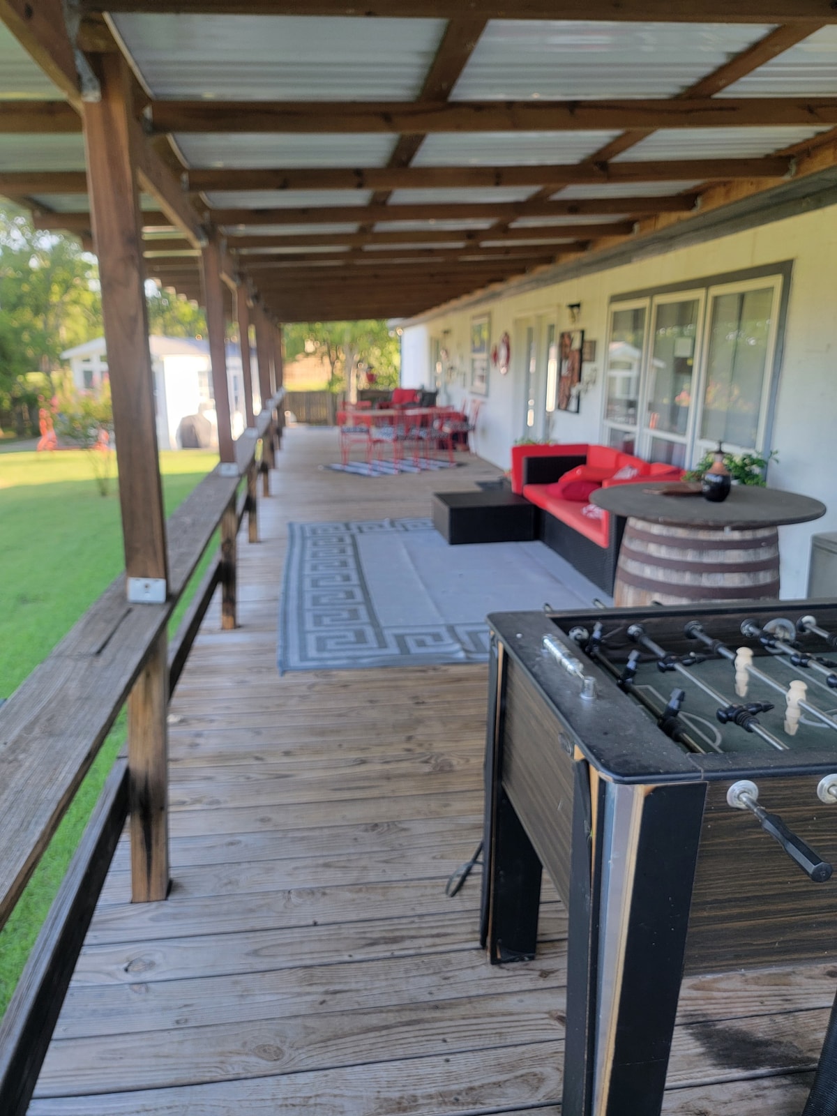 A wooden porch is lined with comfortable seating options, including red sofas and a circular table. A foosball table is positioned in the foreground. A textured area rug complements the outdoor space, which is surrounded by greenery and leads to an open yard.