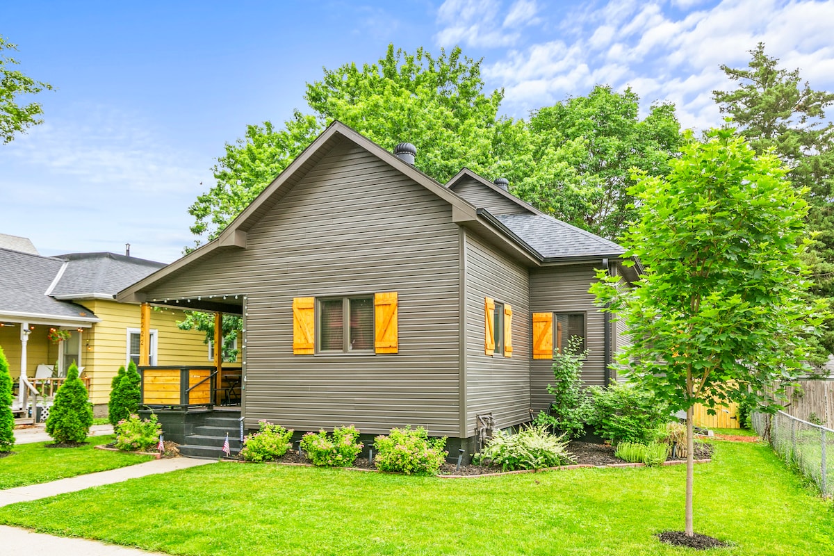 A charming farmhouse exterior is framed by lush green grass and decorative trees. The house features a dark gray facade with vibrant orange shutters, large windows, and a welcoming porch that enhances accessibility to outdoor spaces.