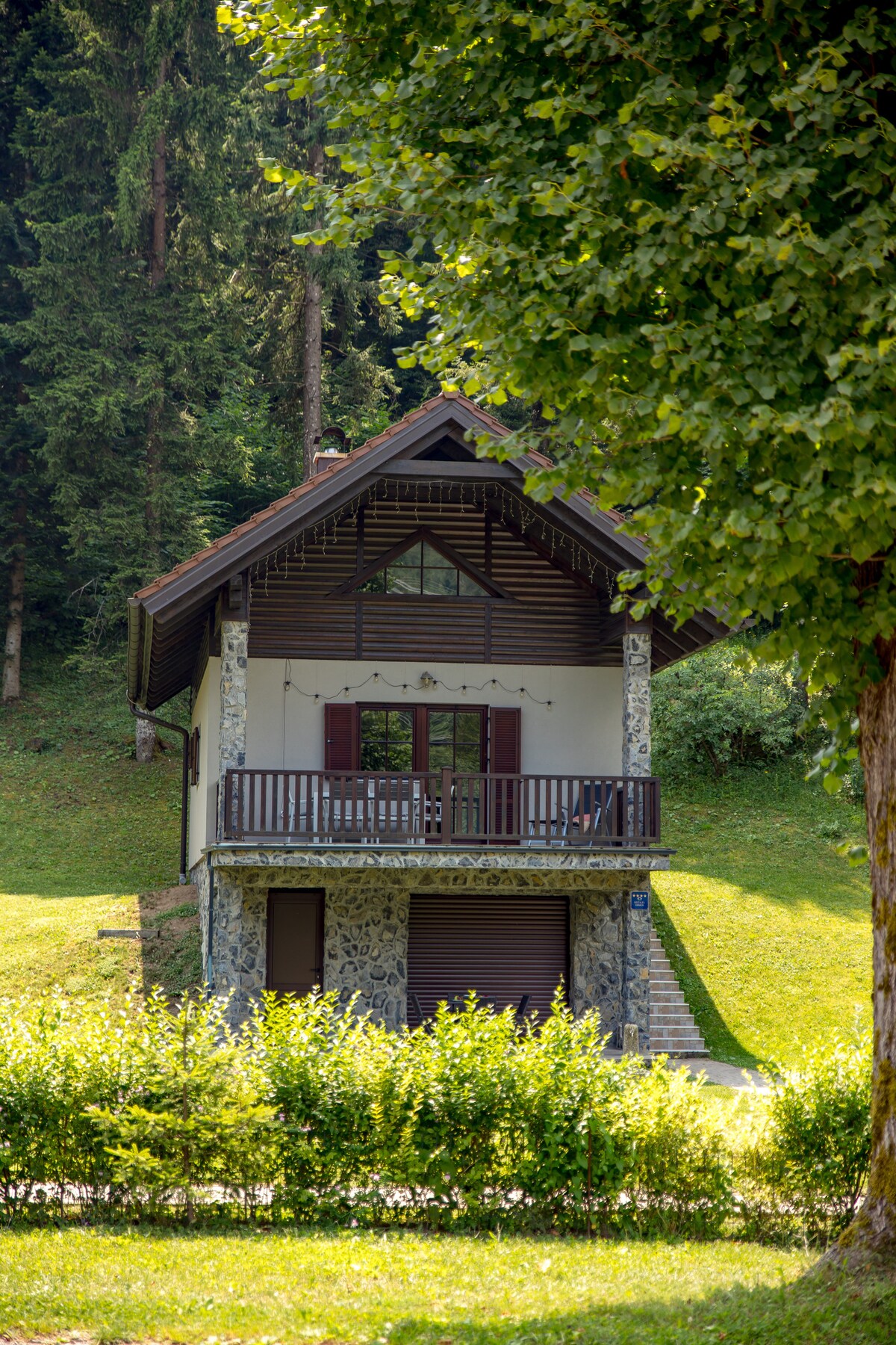 A charming chalet is set amidst a wooded landscape, featuring a stone facade and a covered balcony with wooden railings. The surrounding grassy area is dotted with trees, adding to the serene natural environment.
