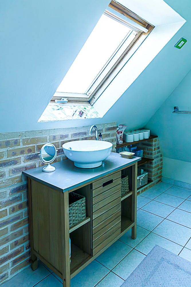A modern bathroom is highlighted by a wooden vanity featuring a round basin and storage baskets. A skylight allows natural light to fill the space, accentuating the pale blue walls and tiled floor. A neatly arranged towel rack and toiletries are visible in the background.