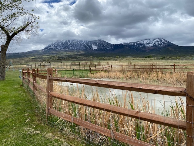 A scenic view of a fenced pond is presented, surrounded by lush greenery and tall grasses. The backdrop features majestic mountains, partially covered in snow, under a cloudy sky. The tranquil landscape embodies the peaceful rural setting.