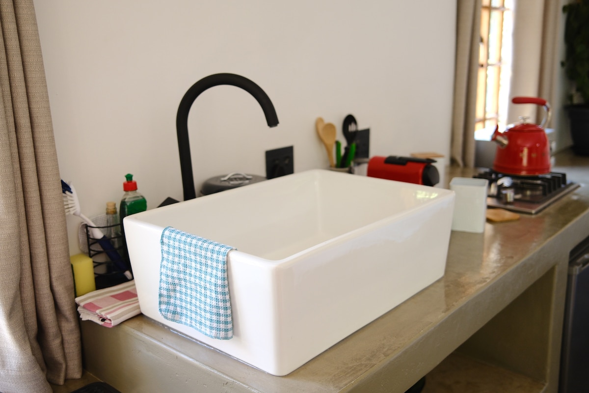 A spacious white sink is positioned on a neutral-toned countertop, complemented by a sleek black faucet. A checkered dishcloth rests over the edge of the sink. Various kitchen utensils and condiments are organized on the countertop, showcasing an inviting cooking space.