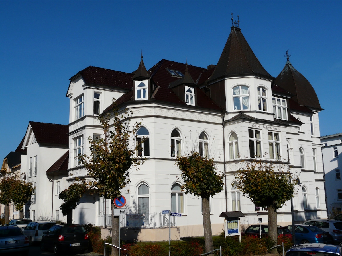 The exterior of a charming historical building is presented, featuring white walls and distinctive architectural elements such as pointed towers and arched windows. Well-maintained trees line the front, adding greenery to the scene. The clear blue sky enhances the building's elegant appearance.