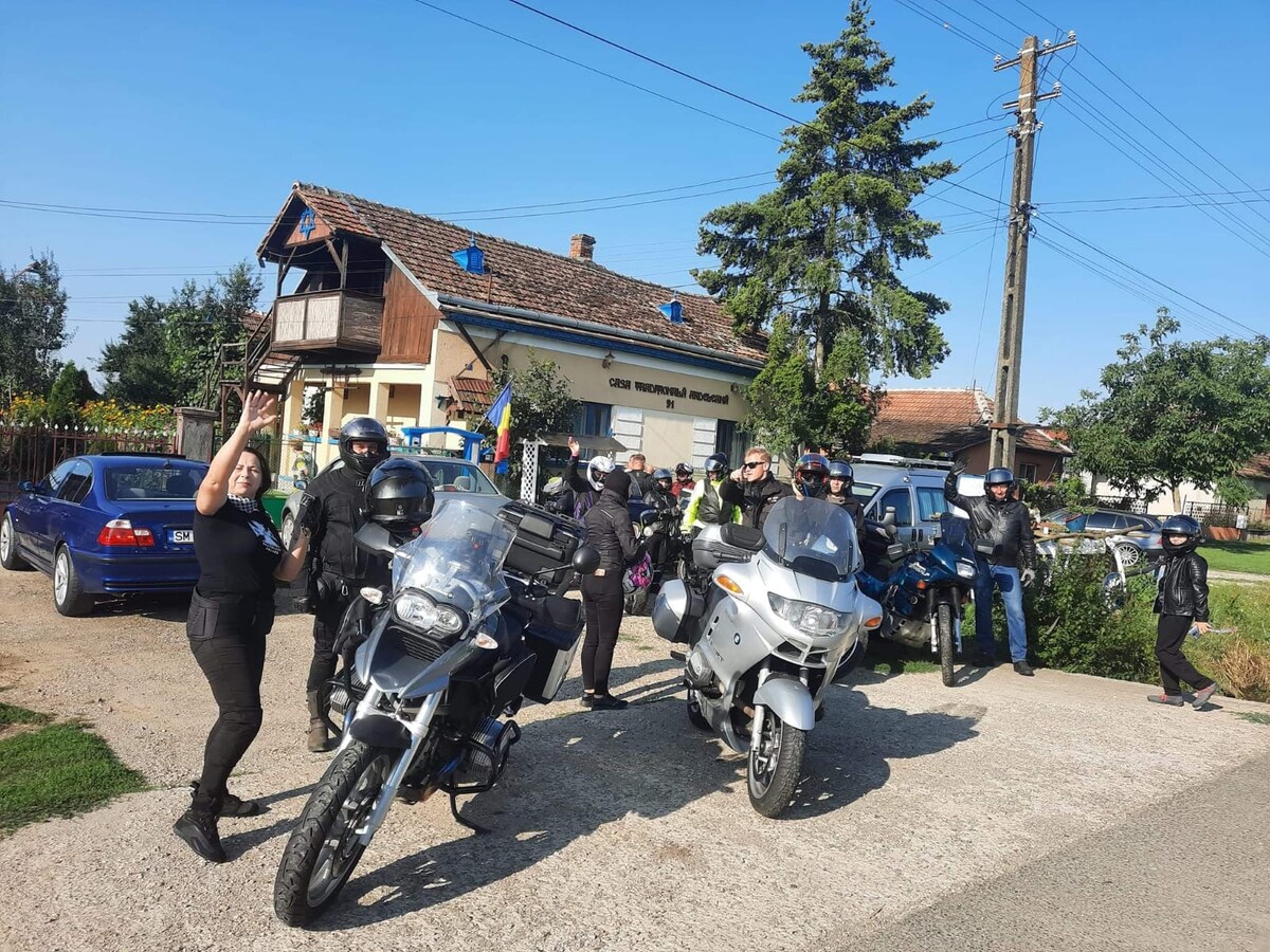 A group of motorcyclists in protective gear congregates outside a rustic building featuring traditional architecture. Several motorcycles are parked nearby, surrounded by vehicles and greenery. The scene captures the camaraderie among riders against a backdrop of a peaceful village setting.