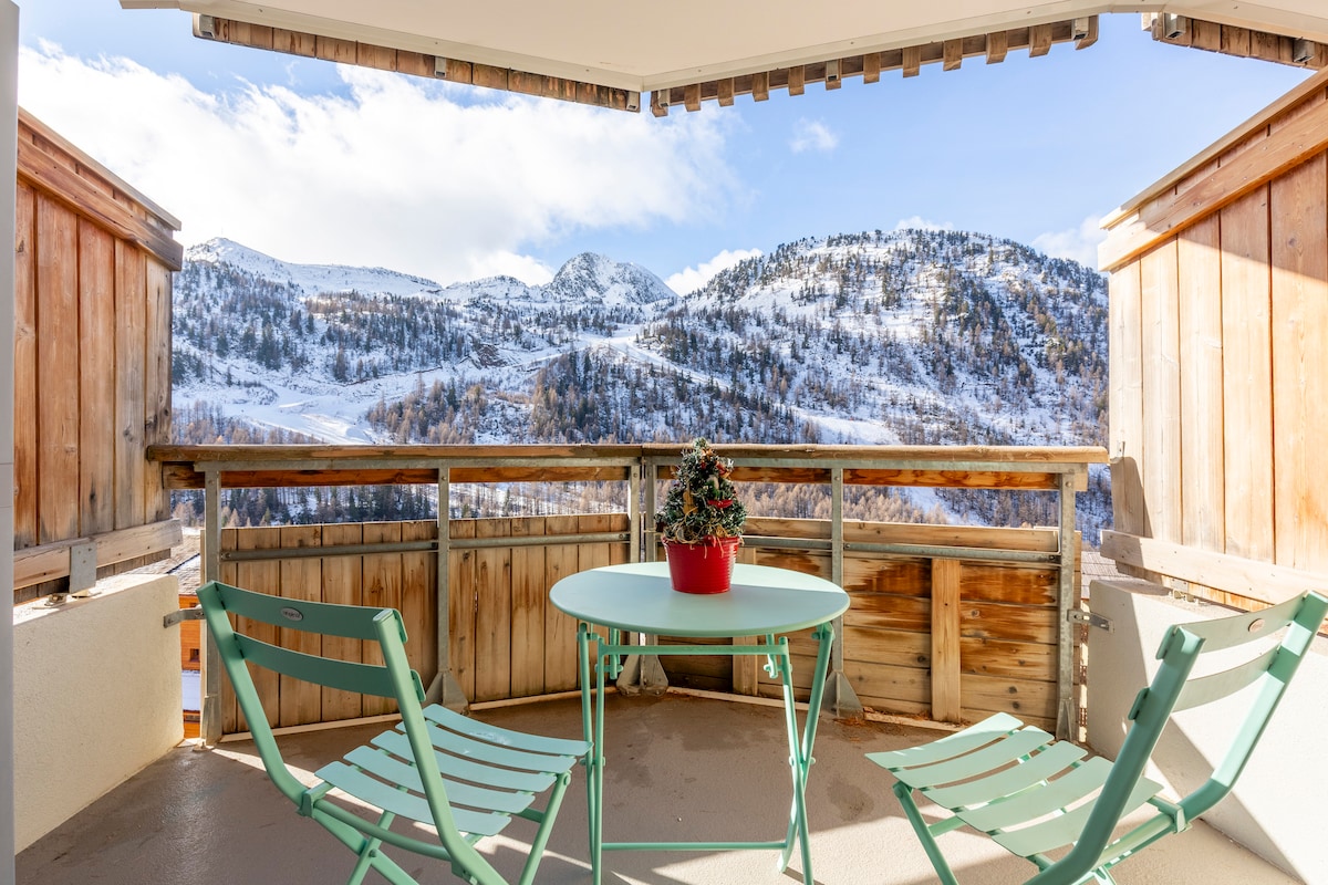 An outdoor terrace features a small round table and two green chairs, offering scenic views of snow-covered mountains and surrounding greenery. Wooden railings surround the space, providing a cozy atmosphere for enjoying the alpine landscape.