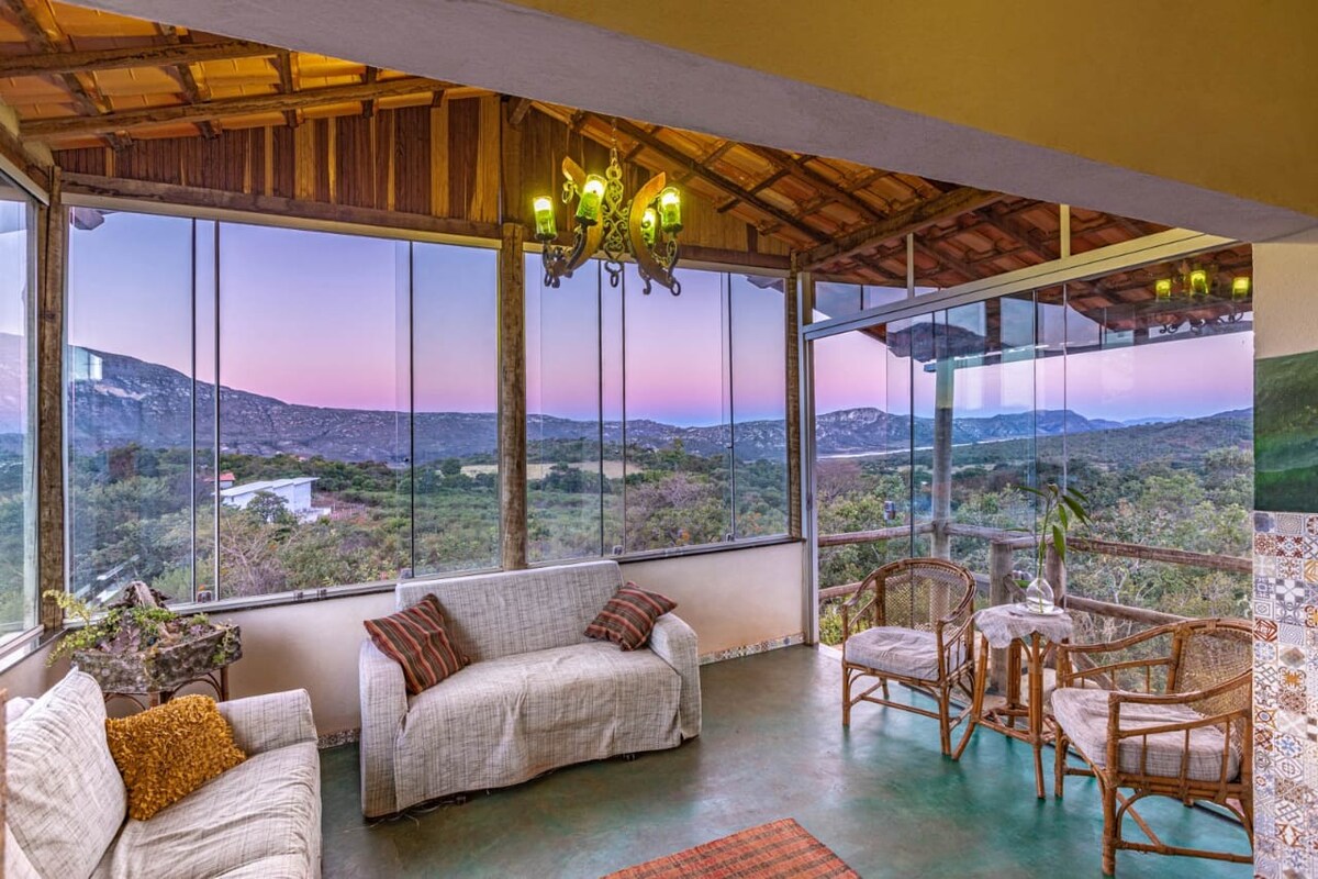 A spacious living area features large glass windows that provide a panoramic view of the surrounding landscape, including distant mountains. Comfortable seating is arranged, with a coffee table in the center. The room is illuminated by a unique chandelier, enhancing the serene atmosphere.