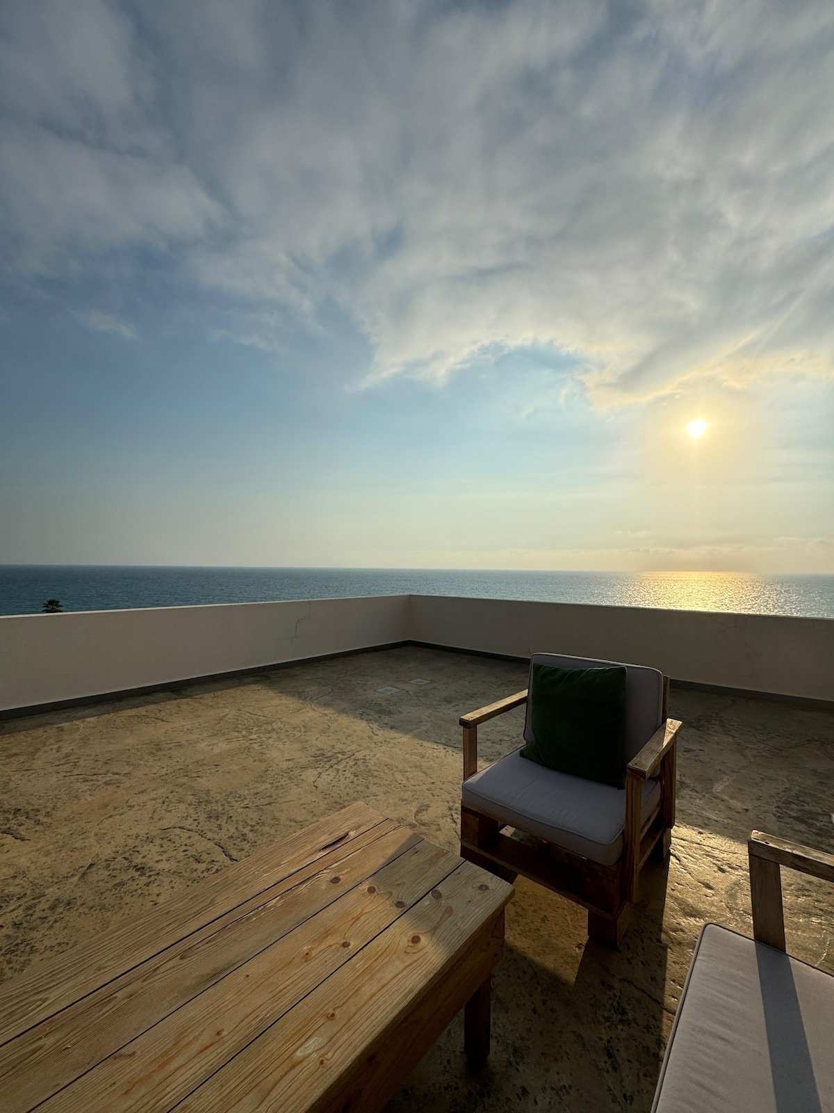 A spacious terrace offers panoramic views of the sea and a sunset sky. Comfortable seating is arranged around a wooden coffee table, complementing the tranquil outdoor space. The serene atmosphere is enhanced by the soft lighting as the sun sets over the horizon.