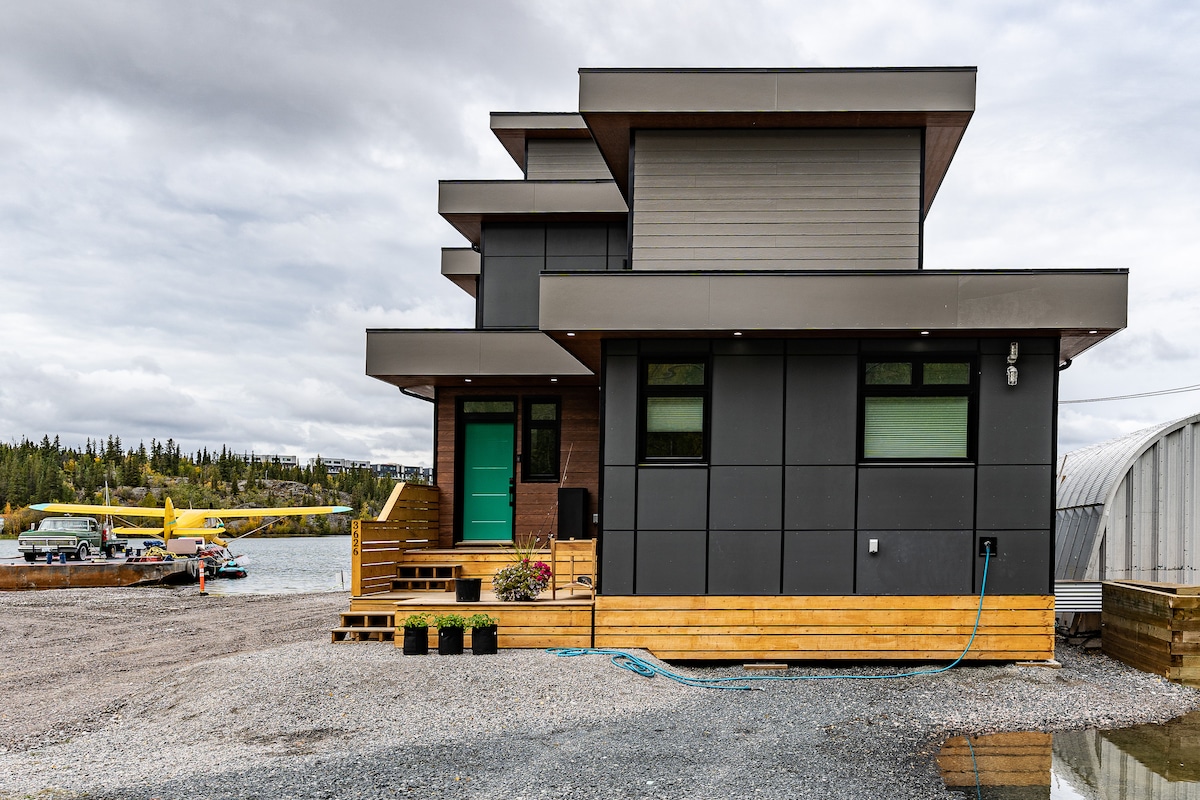 The exterior of the Nordic retreat is modern in design, featuring a combination of dark materials and a distinctive teal door. Surrounded by a gravel yard, the structure offers views of the scenic waterfront and nearby floating planes on Great Slave Lake.