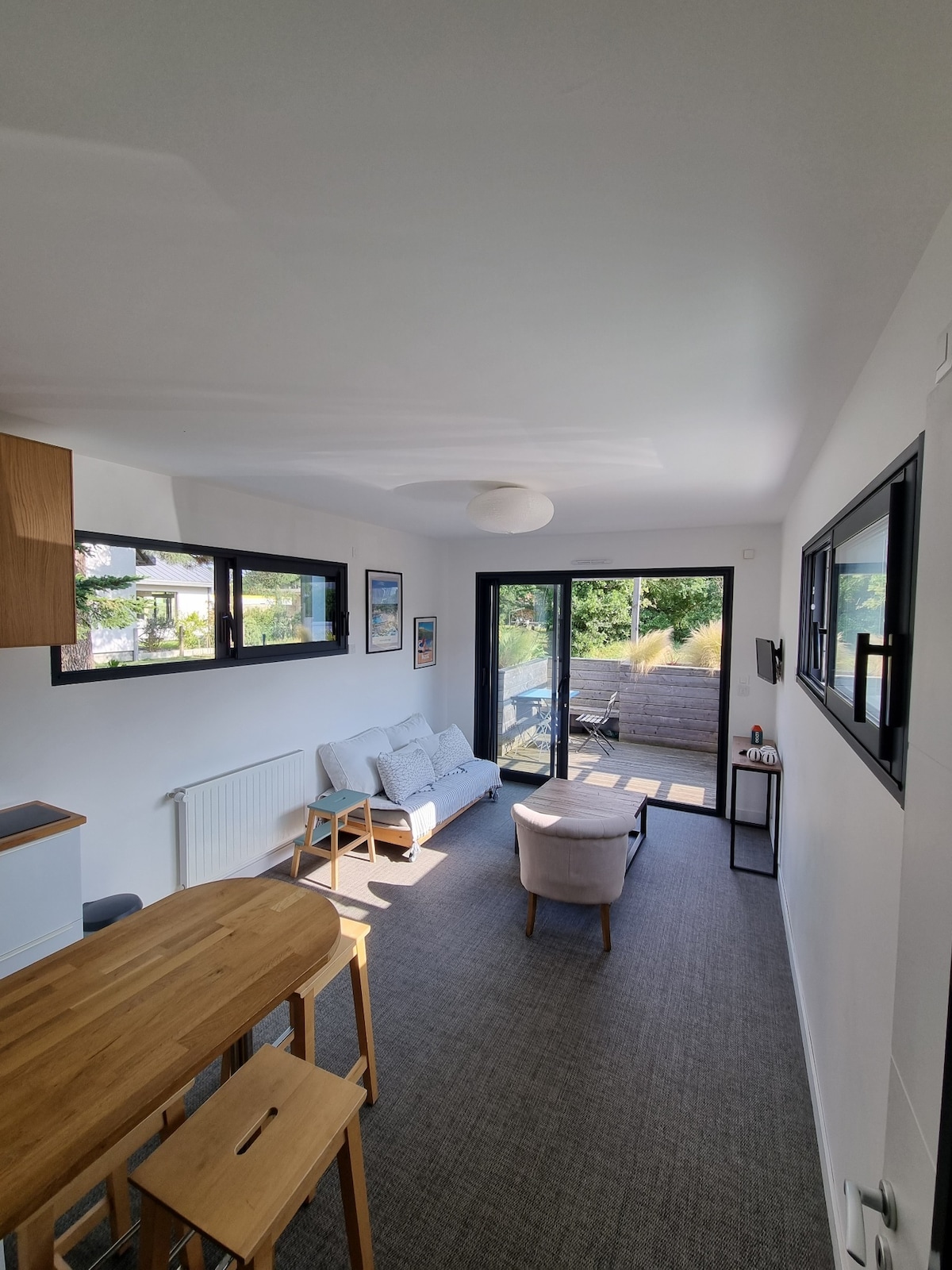 A bright and modern living area is presented, featuring a cozy seating arrangement with a light-colored sofa and a single armchair. Large windows allow plenty of natural light, while a dining table with stools stands nearby, enhancing the open layout.
