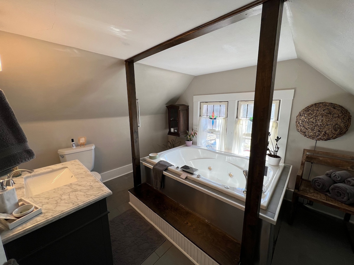 A spacious bathroom features a large jacuzzi tub positioned under sloped ceilings. Two windows allow natural light to illuminate the space, adorned with decorative glass elements. A modern sink with marble countertops and minimalistic decor complements the area, while fluffy towels are neatly arranged nearby.