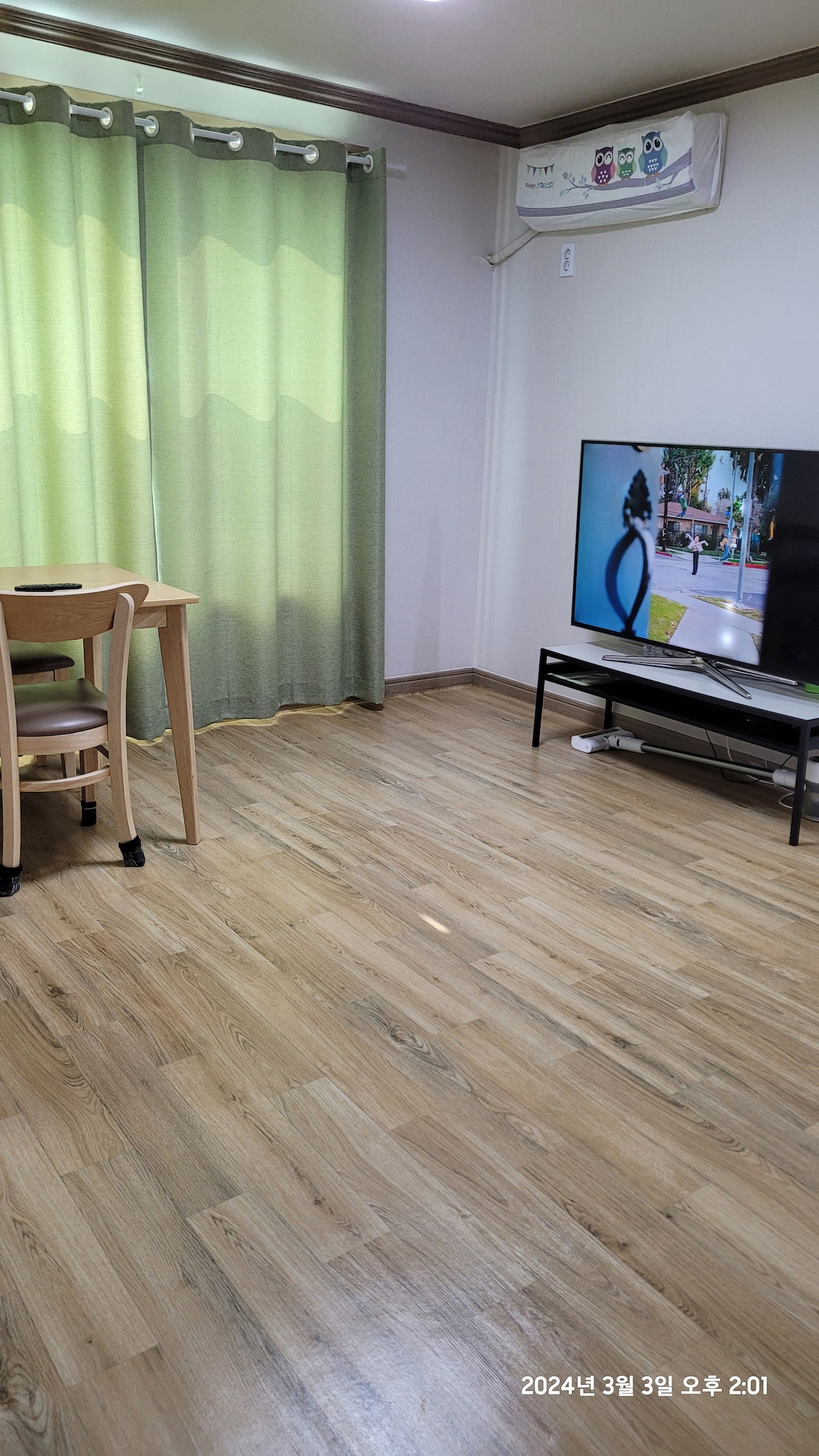 A well-lit interior showcases a small dining table with two chairs beside a window adorned with green curtains. A television on a black stand occupies one corner, while warm wooden flooring enhances the room's inviting feel.
