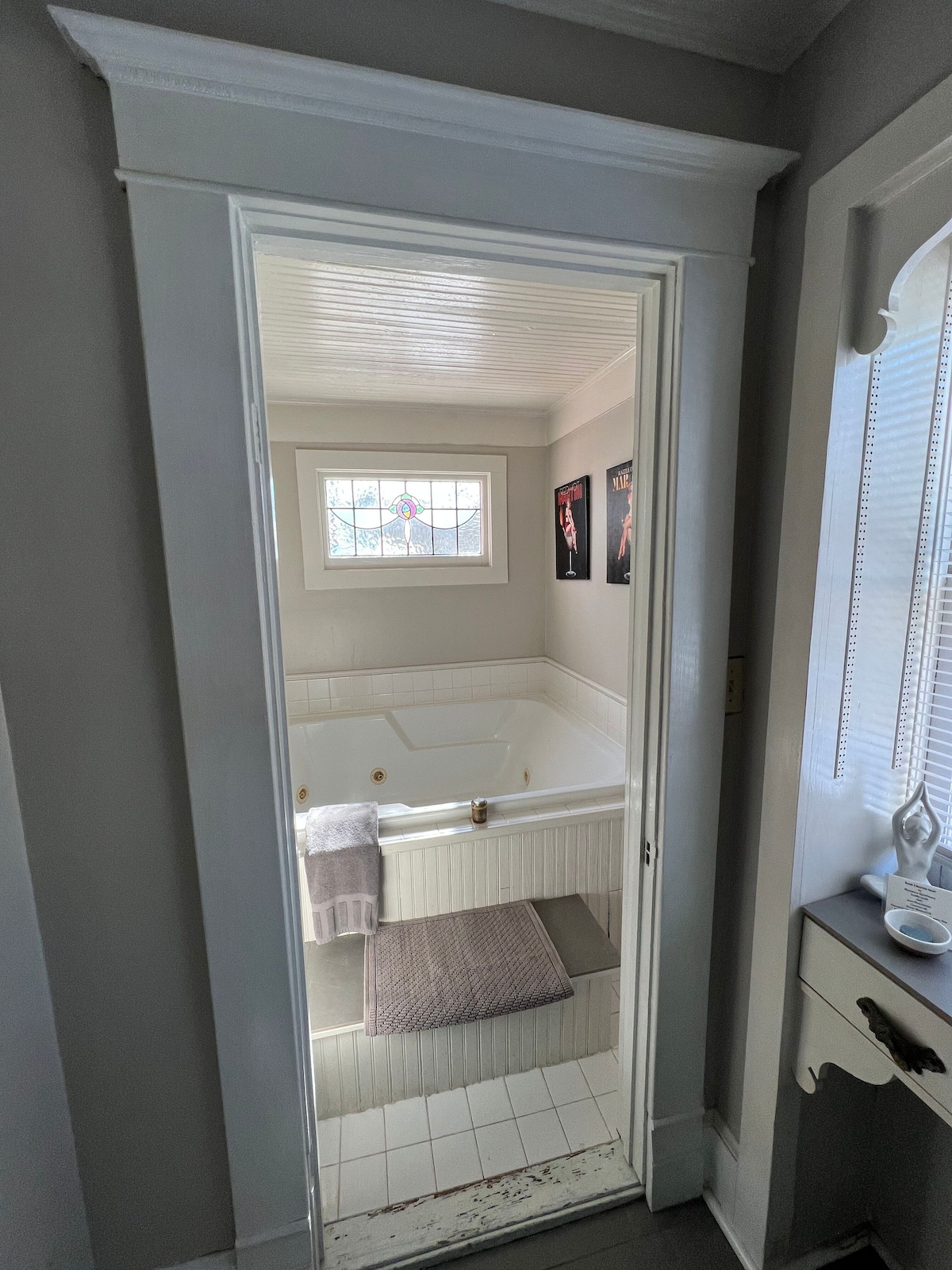 The bathroom entrance features a wide doorway framed in white. Inside, an oversized Jacuzzi tub is visible, complemented by soft towels on a nearby gray rug. Natural light enters through a small window, illuminating the white tiled space decorated with framed wall art.