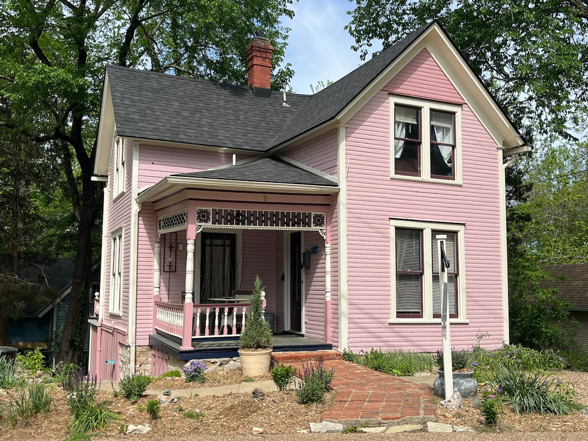 A charming pink Victorian house stands amidst lush greenery, featuring a welcoming front porch with decorative railings and a potted plant. The roof is accented with a classic chimney, and windows adorned with lace curtains reflect a sense of historical elegance.