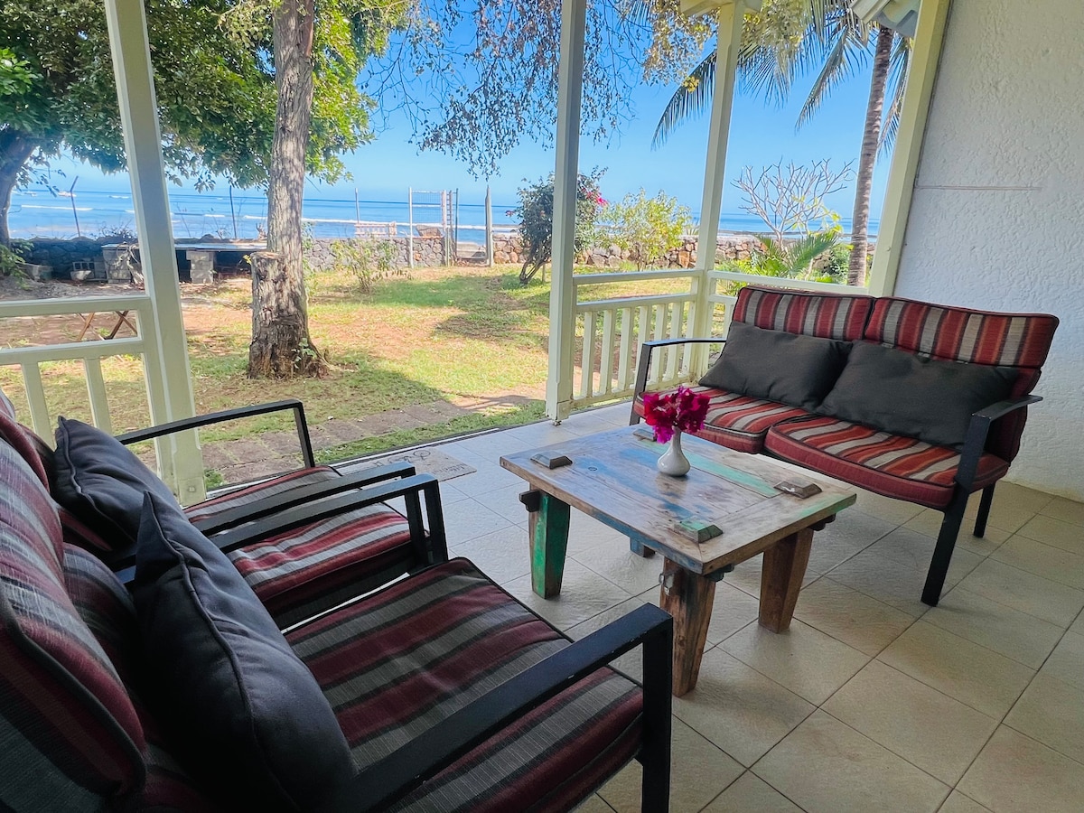 An inviting outdoor seating area features two striped sofas and a rustic coffee table, set against a backdrop of lush greenery. The ocean view can be seen through the open space, enhancing the tranquil atmosphere. Sunlight illuminates the area, creating a relaxed setting.