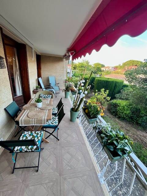 The terrace features a dining table set with a natural fiber placemat, surrounded by folding chairs. Potted plants and flowers are arranged along the railing, contributing to a serene atmosphere. The view includes greenery beyond the terrace, with a hint of blue sky overhead.