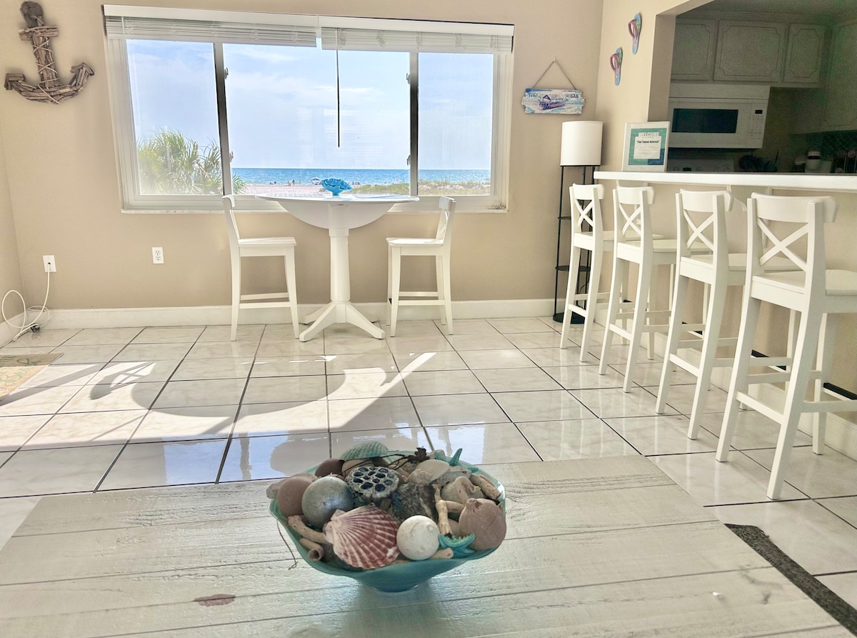 The open living area features a bright view of the beach through a large window. A round dining table is positioned nearby, complemented by a centerpiece of seashells. Bar seating offers additional space, and light tiles reflect the natural ambiance.