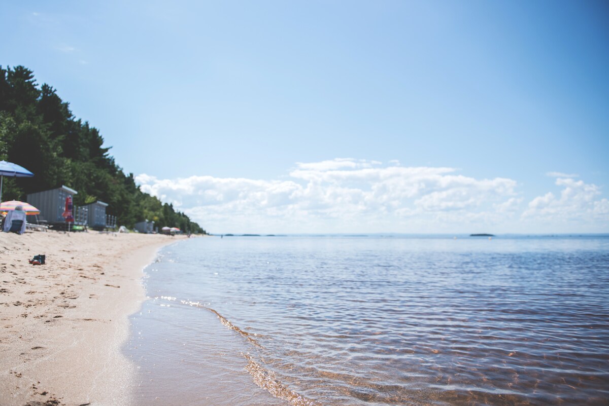 A sandy beach extends along the shoreline, meeting calm waters that reflect the blue sky. Trees line the backdrop, providing a natural setting. A few umbrellas are positioned on the beach, suggesting space for relaxation and enjoying the outdoors.