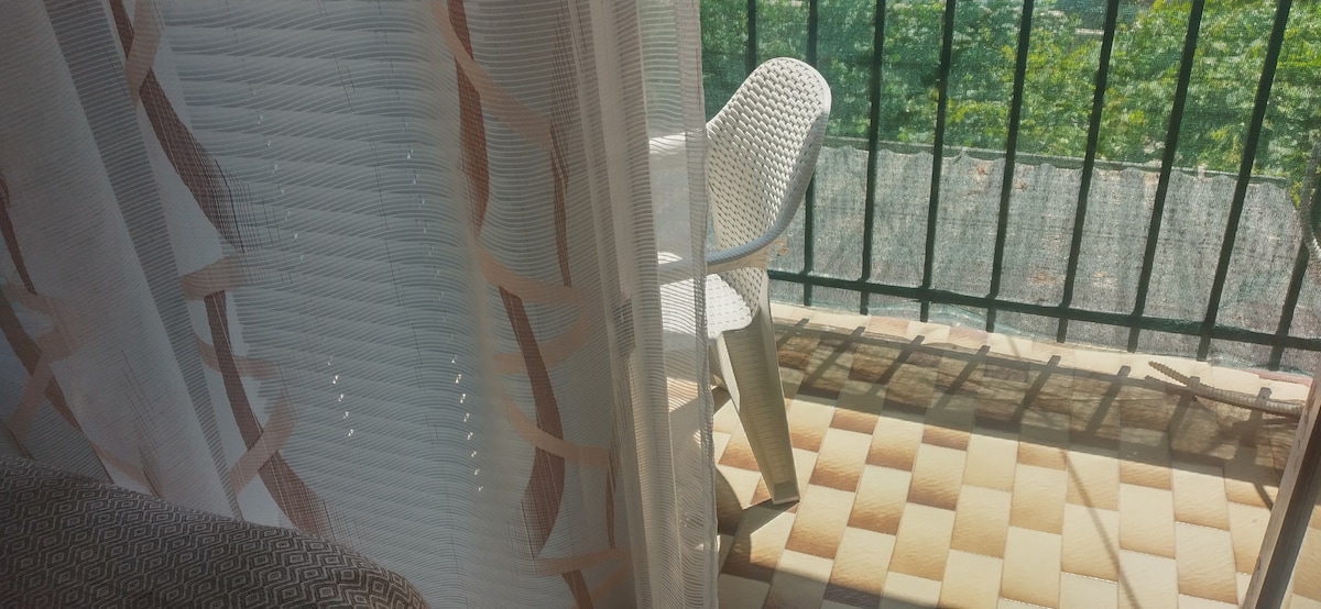 A private balcony is visible, featuring a white chair and a patterned tiled floor. Soft curtains partially filter natural light, accentuating the outdoor space framed by a green railing. Lush greenery can be seen beyond the balcony, creating a serene view.