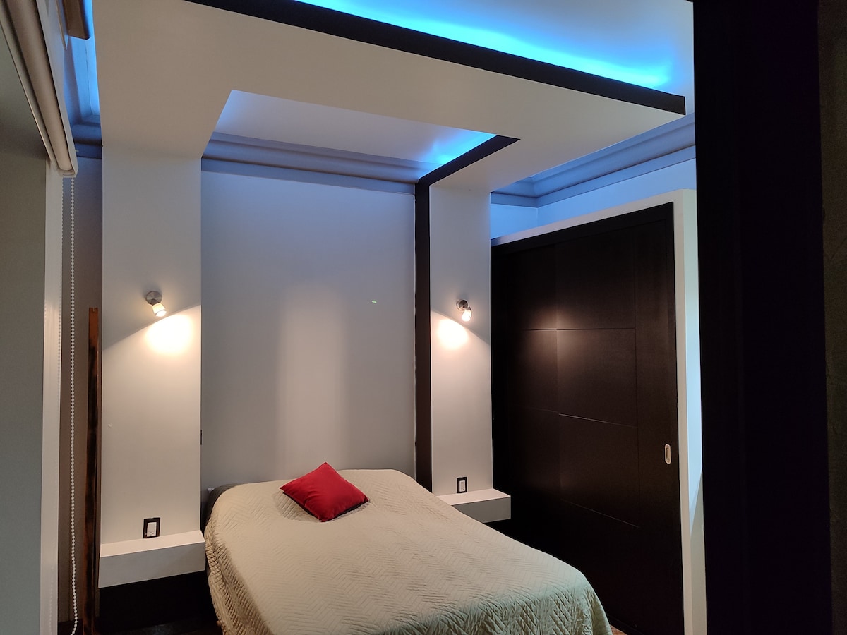 A modern bedroom features a spacious bed with a light quilt and a decorative red pillow. Soft lighting is provided by wall sconces, while ambient blue lighting accents the ceiling. Dark cabinetry and a minimalist design contribute to a sleek and contemporary atmosphere.