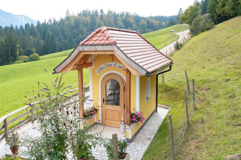 A quaint, yellow structure featuring a wooden exterior and a red-tiled roof is surrounded by lush greenery. Flower beds with vibrant blooms enhance the entryway, while a stone path leads toward the scenic hillside in the background. Trees and mountains provide a tranquil backdrop.