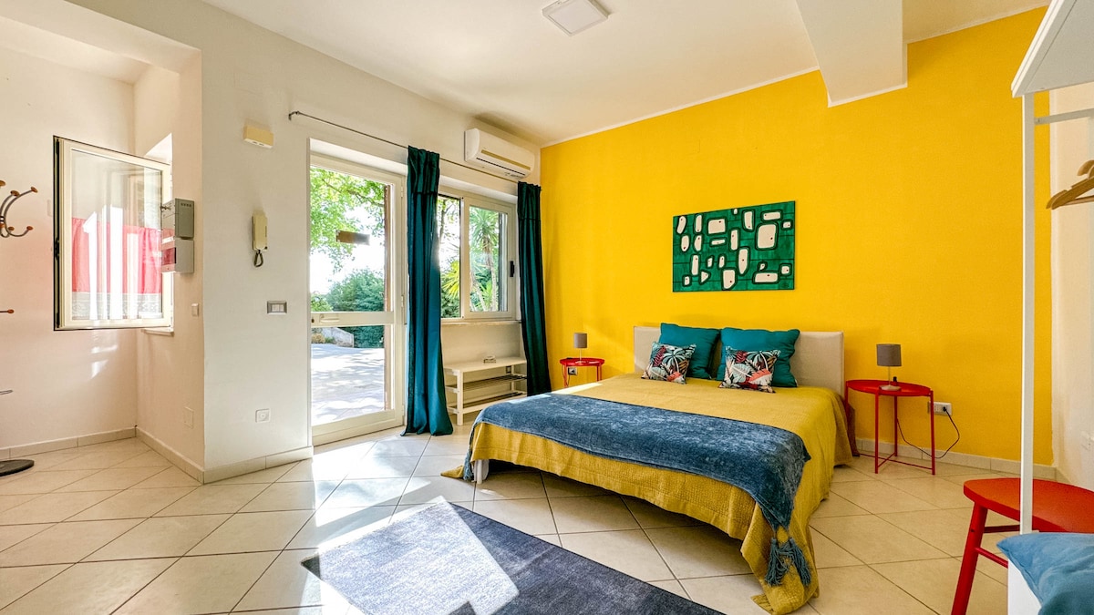 A well-lit sleeping area features a spacious bed with colorful bedding, complemented by decorative cushions. A bright yellow wall adds vibrancy, while large windows offer natural light and views of the outdoor space. A light rug and small side tables enhance the inviting decor.