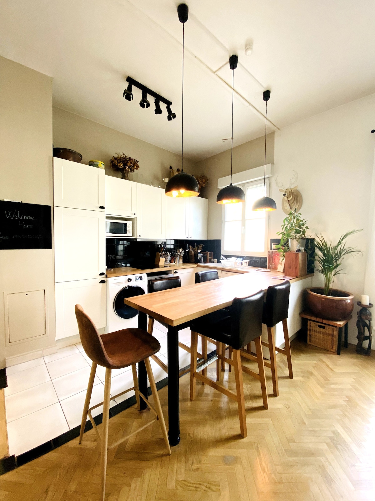 The kitchen features sleek white cabinetry and modern appliances. A wooden dining table with high-backed stools is illuminated by pendant lighting. The space includes a window allowing natural light to enhance the service area, complemented by decorative plants and personal touches.