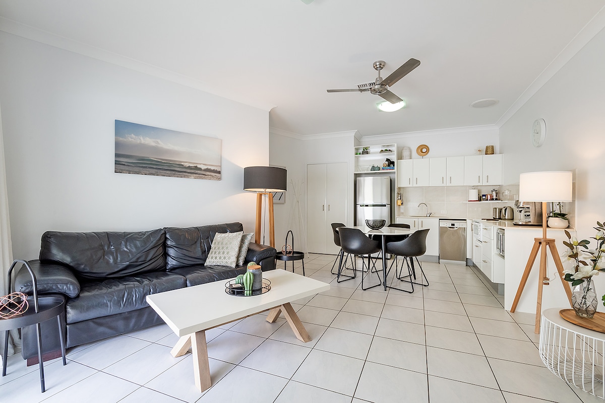 A spacious living area showcases a comfortable leather sofa and a light wood coffee table. A sleek kitchenette with stainless steel appliances is visible. Dining seating for four is arranged nearby. Natural light fills the room, enhancing the clean, modern design.