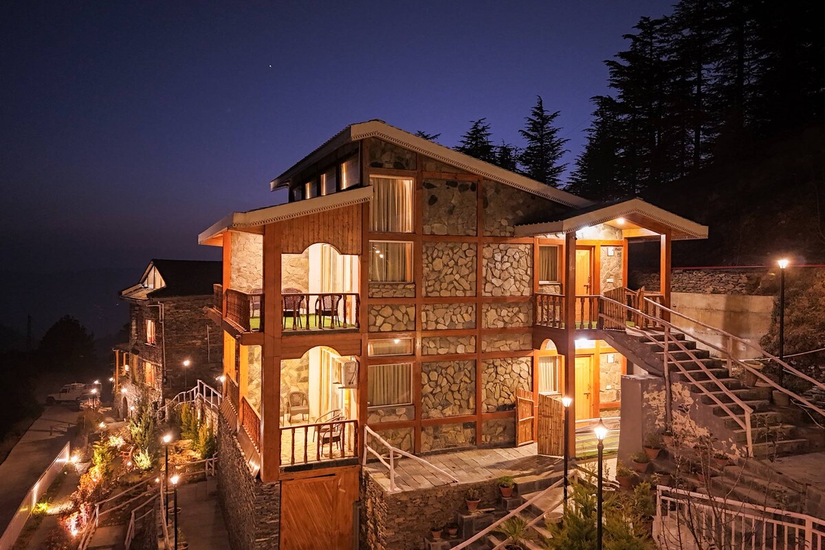 The exterior of Sarion Cottage is captured during twilight, showcasing its traditional stone and wood construction. Soft lighting illuminates the balconies and pathways, creating a welcoming ambiance. Lush deodar trees surround the property, enhancing its serene hilltop location.