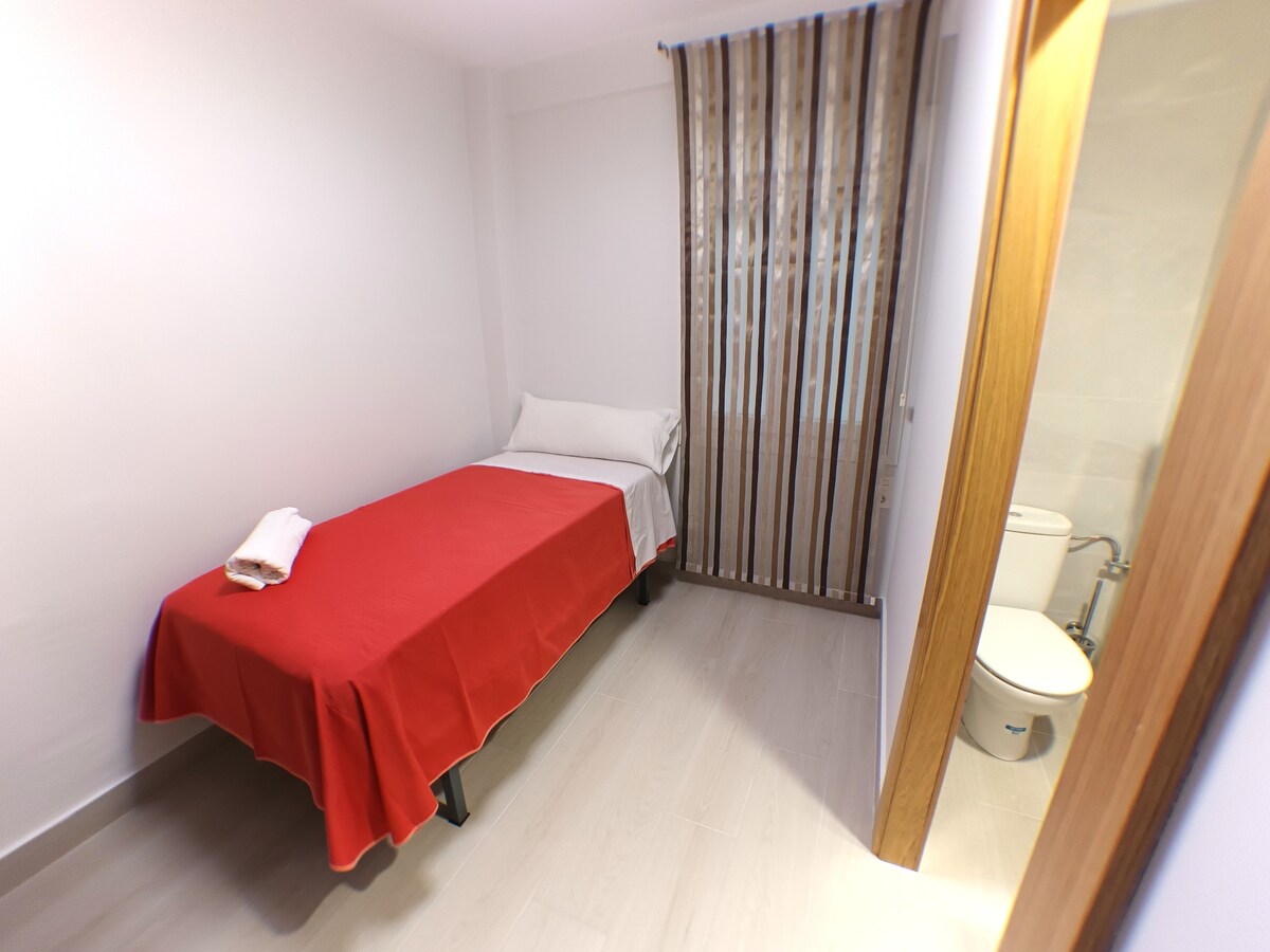 A simple bedroom features a single bed covered with a vibrant red bedspread. A clean towel is neatly placed on the bed. A window with striped curtains allows soft light to enter. A bathroom is visible in the corner, equipped with modern amenities.