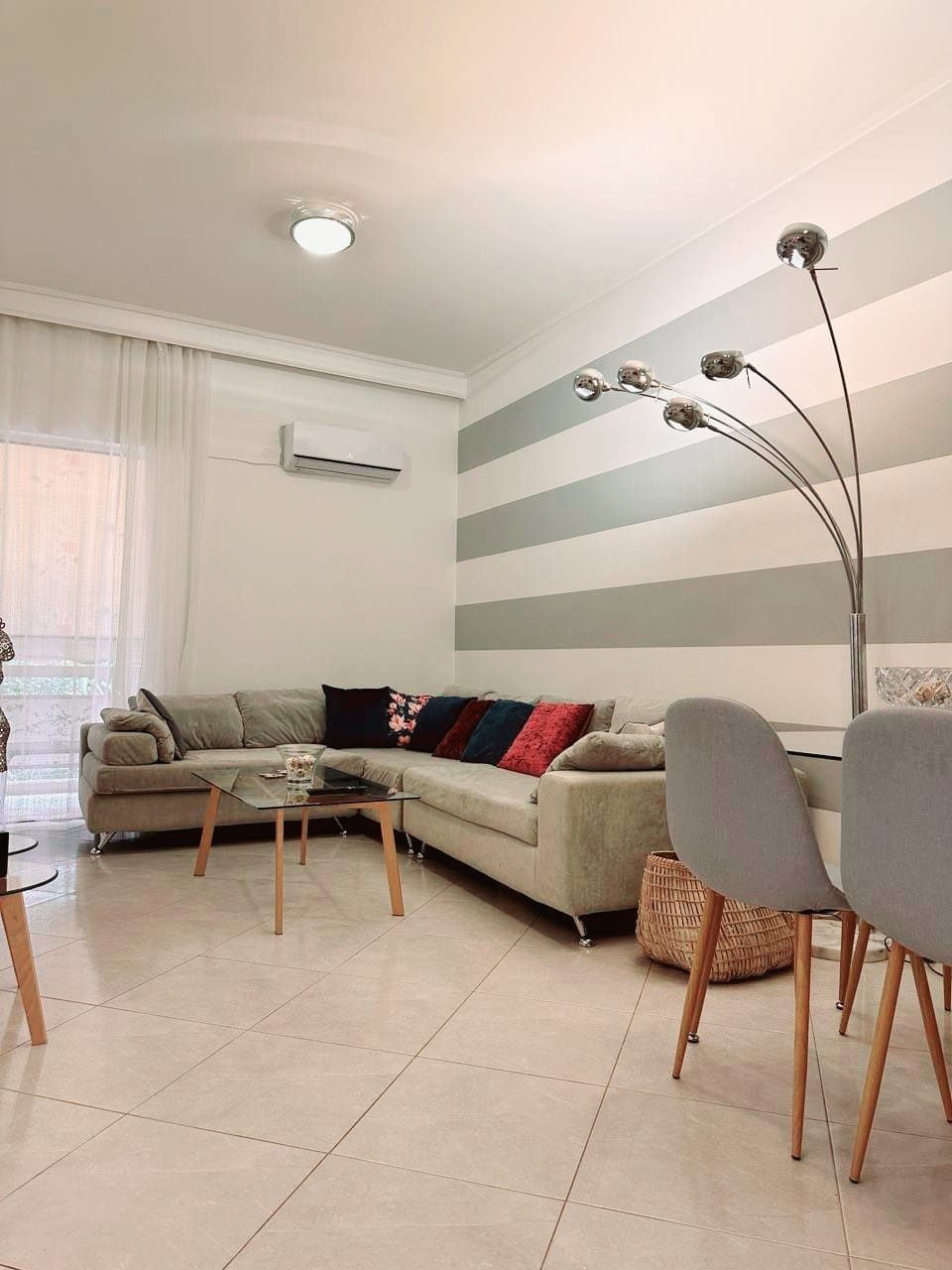 A modern living area features a light-colored sectional sofa adorned with decorative pillows in varied hues. A glass coffee table stands at the center, complemented by sleek dining chairs. Natural light filters through sheer curtains, illuminating the neutral-toned striped wall.