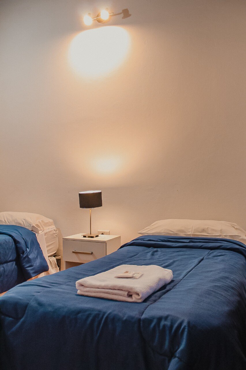 A sleeping area is presented with two individual beds covered in blue bedding. A white towel is neatly arranged on one bed. Beside the beds, a nightstand holds a small lamp that provides soft lighting, illuminating the simple, clean design of the space.