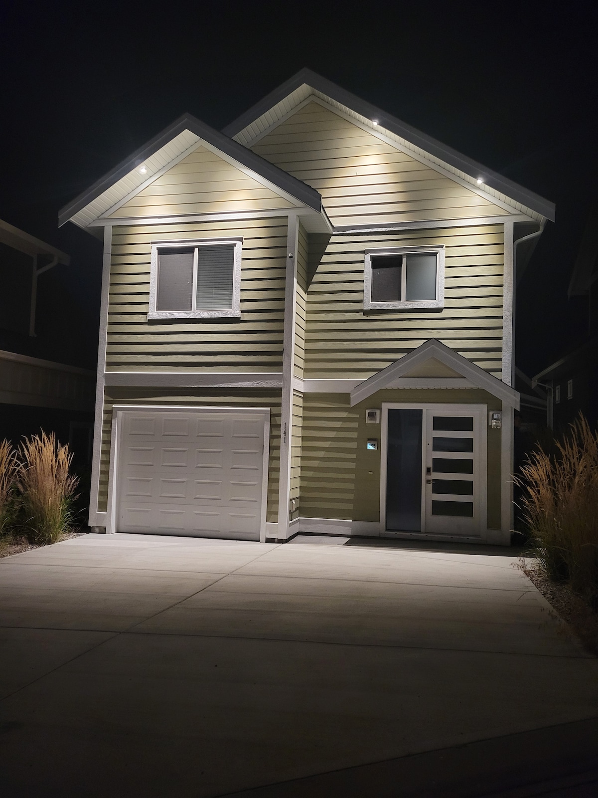 The cottage exterior is illuminated by soft lighting against the night sky. The two-story building features a light green facade with white trim, large windows, and a modern front door. A well-maintained driveway leads up to the garage, framed by ornamental grasses.