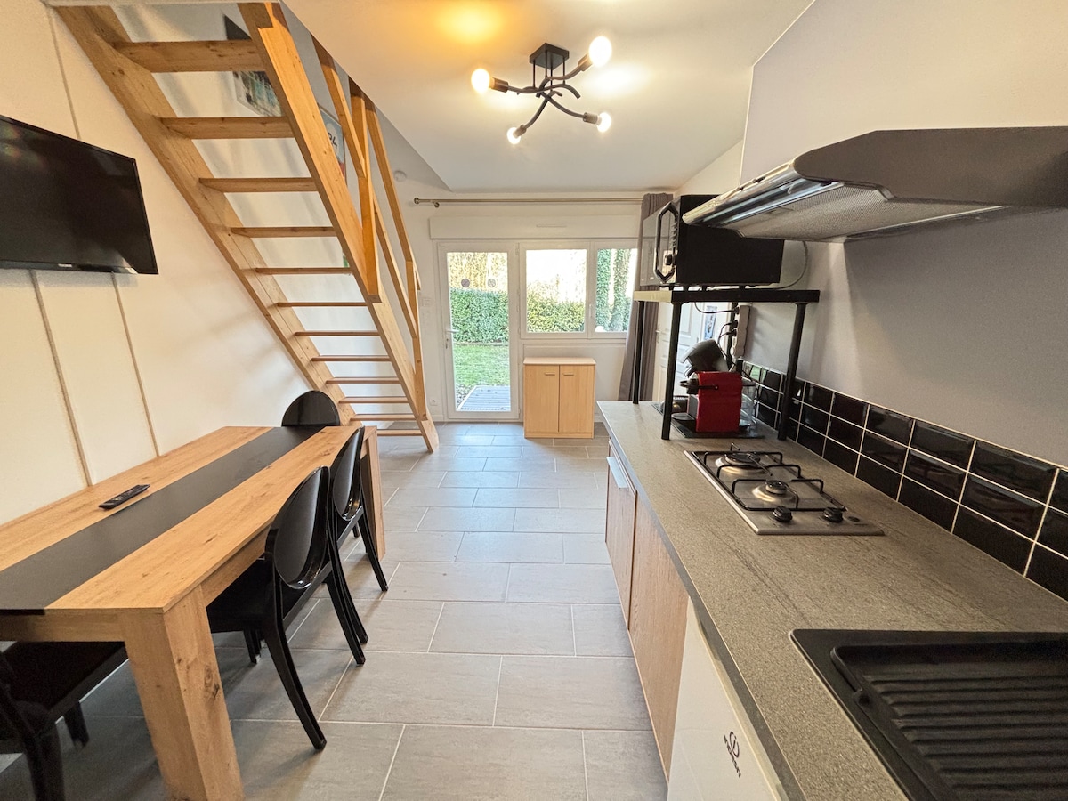 The open living area features a stylish kitchen equipped with modern appliances, including a gas stove. A wooden dining table is accompanied by black chairs, and a staircase leads to the upper level. Large glass doors provide a view of the garden and allow natural light to enter.