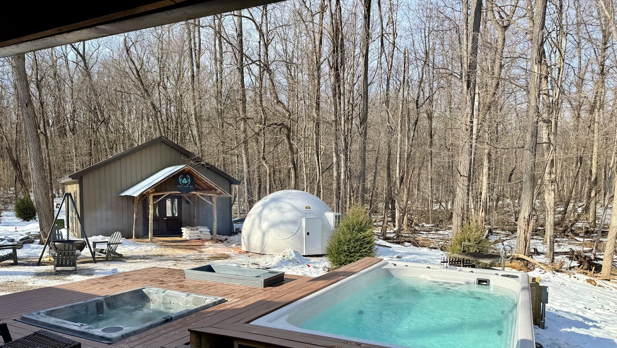 Two hot tubs are nestled on a wooden deck, surrounded by trees in a serene winter landscape. A structure resembling a dome is visible in the background, alongside a rustic building. Snow blankets the ground, enhancing the peaceful atmosphere.