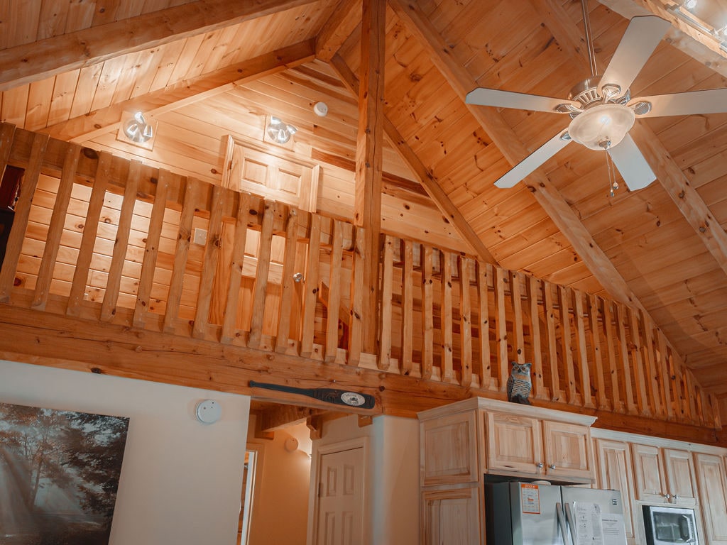 The interior of the cabin is characterized by wooden beams and a vaulted ceiling, creating a spacious and inviting atmosphere. A loft area is visible above, with a wooden railing that adds to the rustic charm. Bright ceiling lights enhance the warm tones of the wood.
