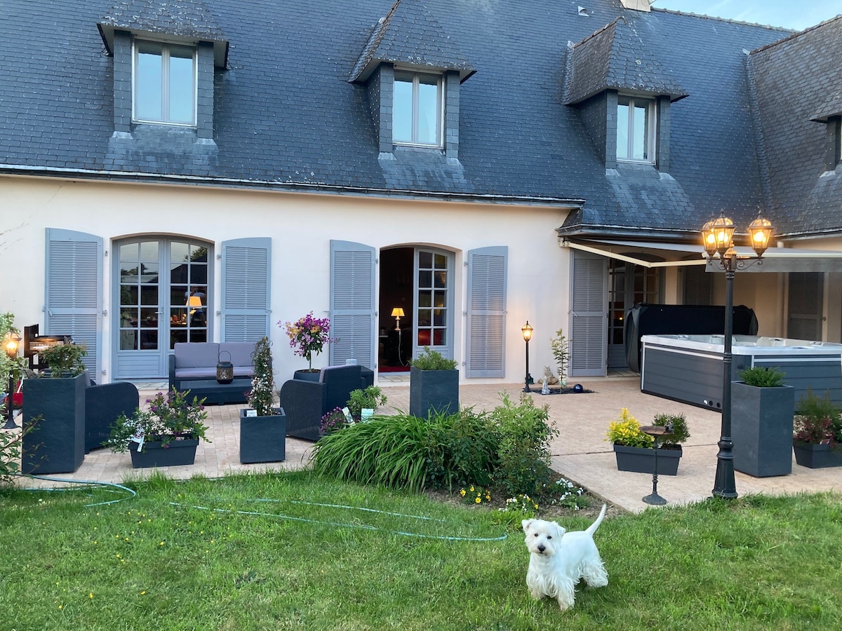 The exterior of the apartment is showcased, featuring a spacious terrace with potted plants and comfortable seating. A jacuzzi is situated nearby, with decorative lamps adding a warm touch. A small, white dog is playfully positioned on the grass, providing a lively element to the tranquil setting.