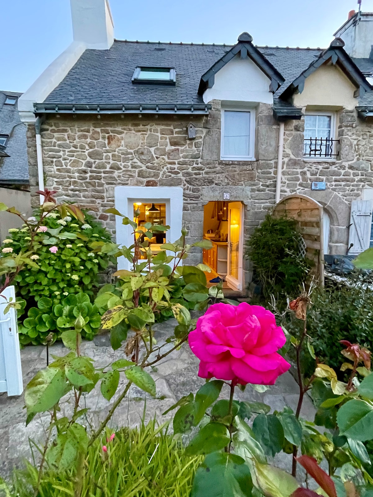 A charming stone cottage is framed by vibrant greenery and a pink rose bush in the foreground. The entrance is illuminated, hinting at a warm interior. The cottage features a slate roof and subtle architectural details, blending harmoniously with the surrounding landscape.