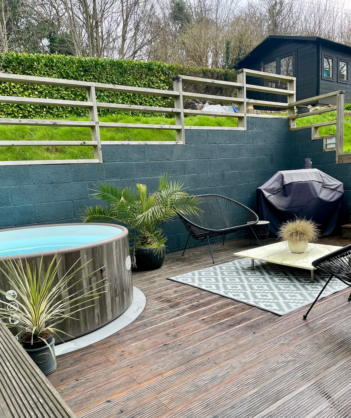 A wooden hot tub is situated on a wooden deck surrounded by lush greenery. Black chairs and a small table with a decorative plant enhance the outdoor space. A charcoal grill is partially covered, providing options for outdoor dining and relaxation.