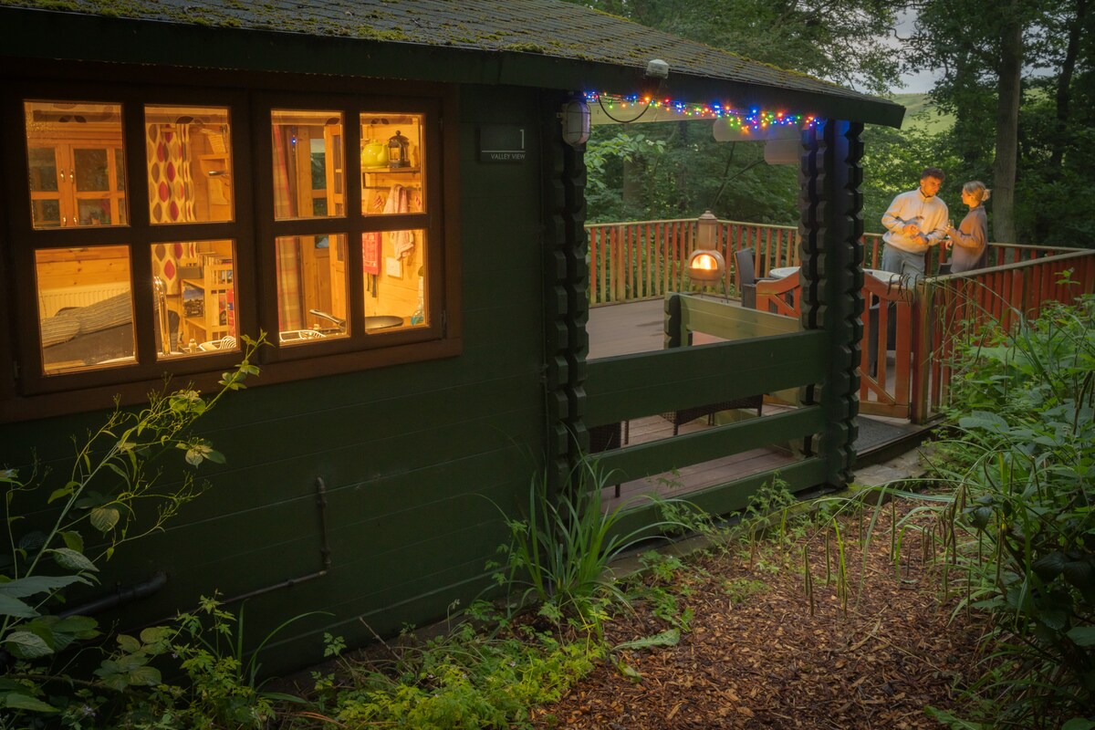 The exterior of the log cabin is framed by lush greenery, showcasing a spacious terrace with fairy lights. Dim interior lighting highlights a welcoming atmosphere. The cabin features multiple windows, allowing natural light to fill the space, creating an inviting setting for guests.