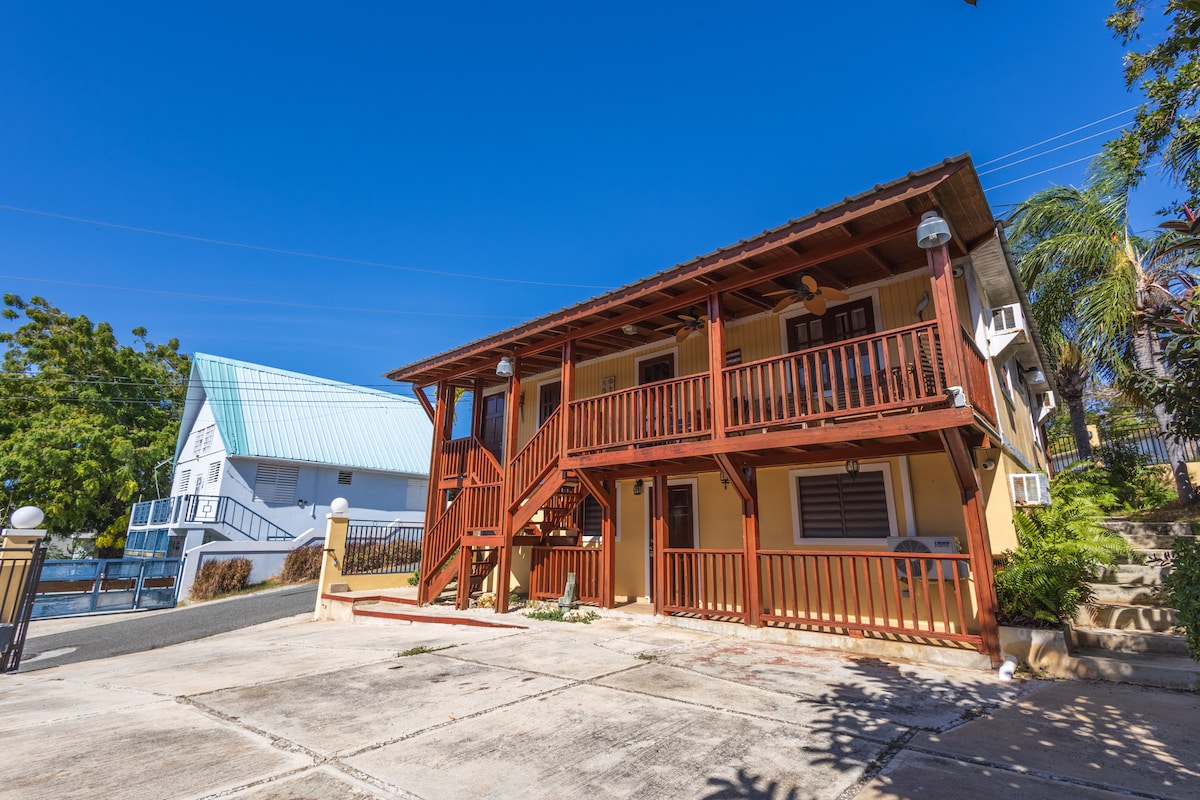 A two-story apartment building features a warm orange exterior with wooden balconies and stairs. Surrounded by greenery, the concrete parking area is visible in front, along with a neighboring building with a blue roof. Clear blue skies create a bright atmosphere.