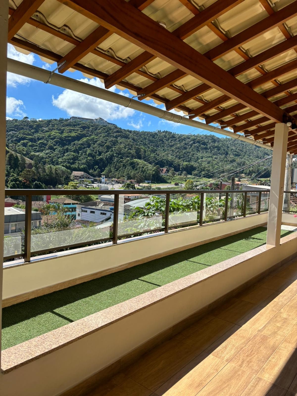 A spacious outdoor balcony is shown, featuring a clear glass railing that offers unobstructed views of the surrounding hills and greenery. A narrow green area runs along the base, while the balcony floor is covered with light-colored wood planks. Sunlight brightens the scene.