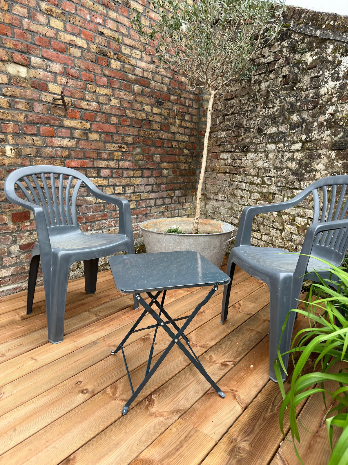 A small outdoor seating area features two gray plastic chairs positioned around a foldable metal table. A copper planter with a small tree is centered in the space, surrounded by the natural wood decking and a brick wall, creating a cozy atmosphere.